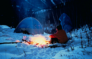 A person in a red jacket sits by a fire outside a tent in a snowy forest during twilight, capturing the serene and chilly ambiance of winter camping.
