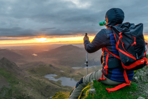 A hiker seated on a mountain edge at sunset, overlooking a valley with lakes, holding two trekking poles, and wearing a backpack.