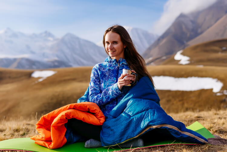 A woman in a blue jacket wrapped in a comfortable sleeping bag sits on a yoga mat in a mountainous landscape, holding a cup and smiling, with snowy peaks in the background.