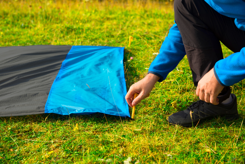 A person in a blue jacket and black shoes secures the corner of the best tent footprint to the grass with a metal peg.