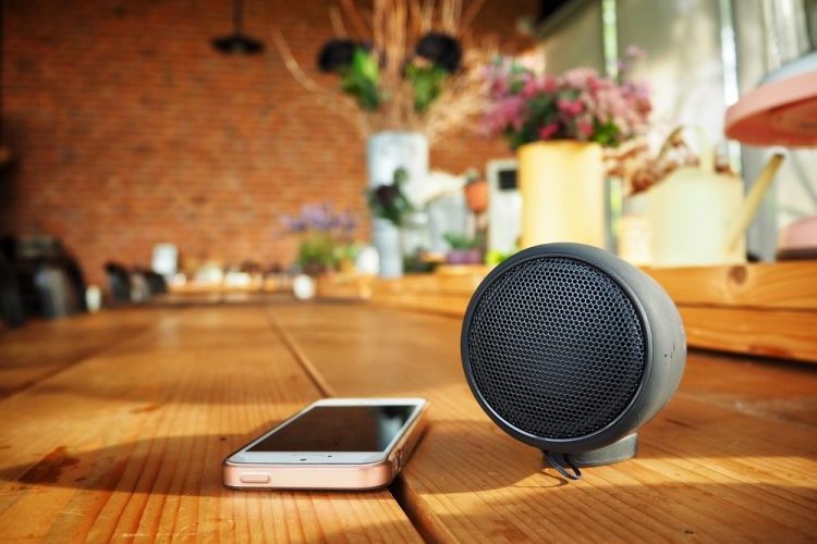 A portable round Bluetooth speaker next to a smartphone on a wooden table, inside a cozy café with soft lighting and vibrant flowers in the background.