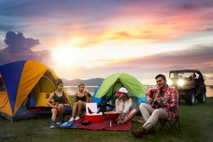 Group of friends enjoying a camping trip by a lake at sunset, with one playing guitar by colorful tents. Vibrant sky and natural scenery in the background, some friends sleeping in their car at the campground.