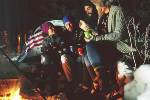 Four friends enjoying a campfire in a snowy forest at night, holding mugs and engaged in conversation about what to pack for winter camping. They're warmly dressed in winter hats and jackets.