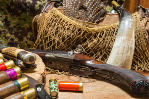 A collection of adaptive vintage hunting gear, including a flintlock rifle, ammunition, a hunting knife, and a camouflage jacket, arranged on a wooden surface.