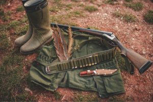 A hunter's adaptive gear laid on the ground, featuring green rubber boots, a shotgun, a belt with cartridges, a green jacket, and a knife with a wooden handle.