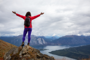 A person with arms spread wide stands on a mountain peak overlooking a majestic lake and mountain landscape under cloudy skies, embodying the spirit of adaptive hiking.