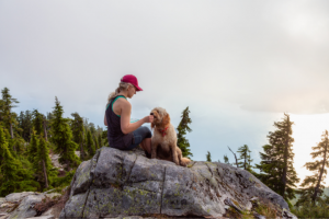 A woman in a tank top and hat sits on a rocky peak with a fluffy dog, engaging in adaptive hiking, surrounded by forested mountains under a cloudy sky.