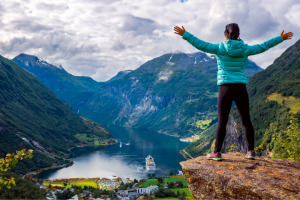 A person in a turquoise jacket and black pants stands with arms outstretched on a cliff overlooking a scenic fjord with a cruise ship and small village below, engaging in adaptive hiking surrounded by lush green mountains.