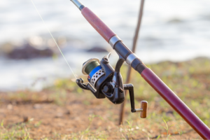 Close-up of the best fishing pole holder for wheelchair users with a reel, propped up on a shoreline with a blurred background featuring water and rocks.