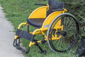 A yellow power wheelchair with black accents is parked outdoors on a dirt path next to some green shrubbery.