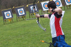 A young archer in a red, white, and black uniform using a compound bow with draw loc disabled archery equipment to aim at a target in a grassy field lined with multiple archery targets.